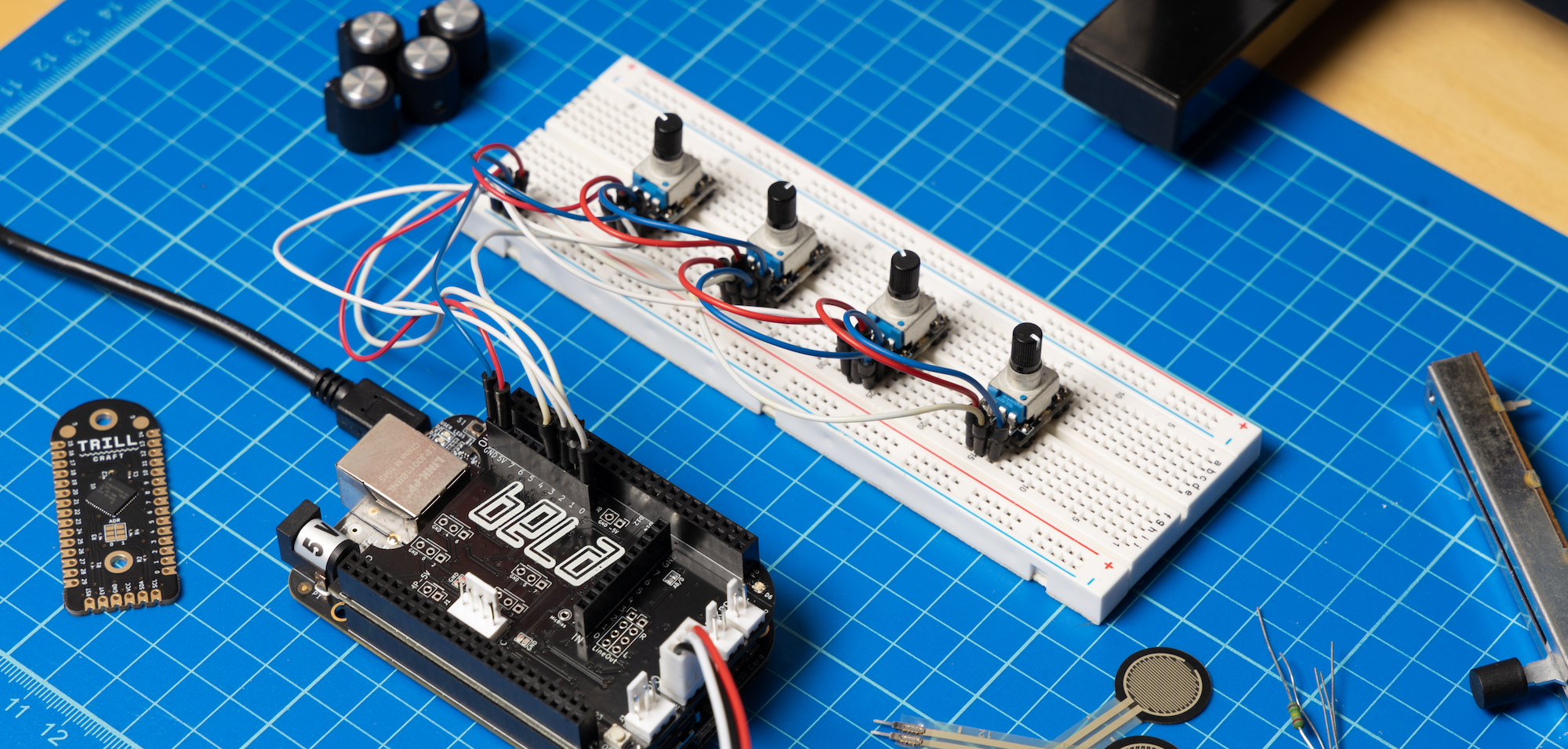 Hands working with a Bela system on a lab bench, surrounded by electronic parts and tools