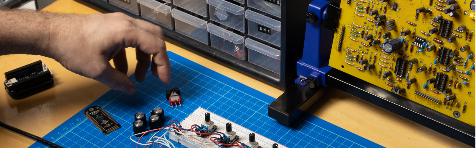 Hands working with a Bela system on a lab bench, surrounded by electronic parts and tools
