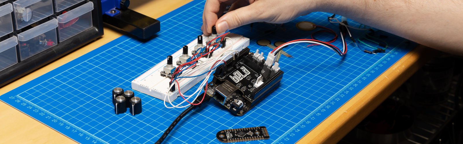 Hands working with a Bela system on a lab bench, surrounded by electronic parts and tools